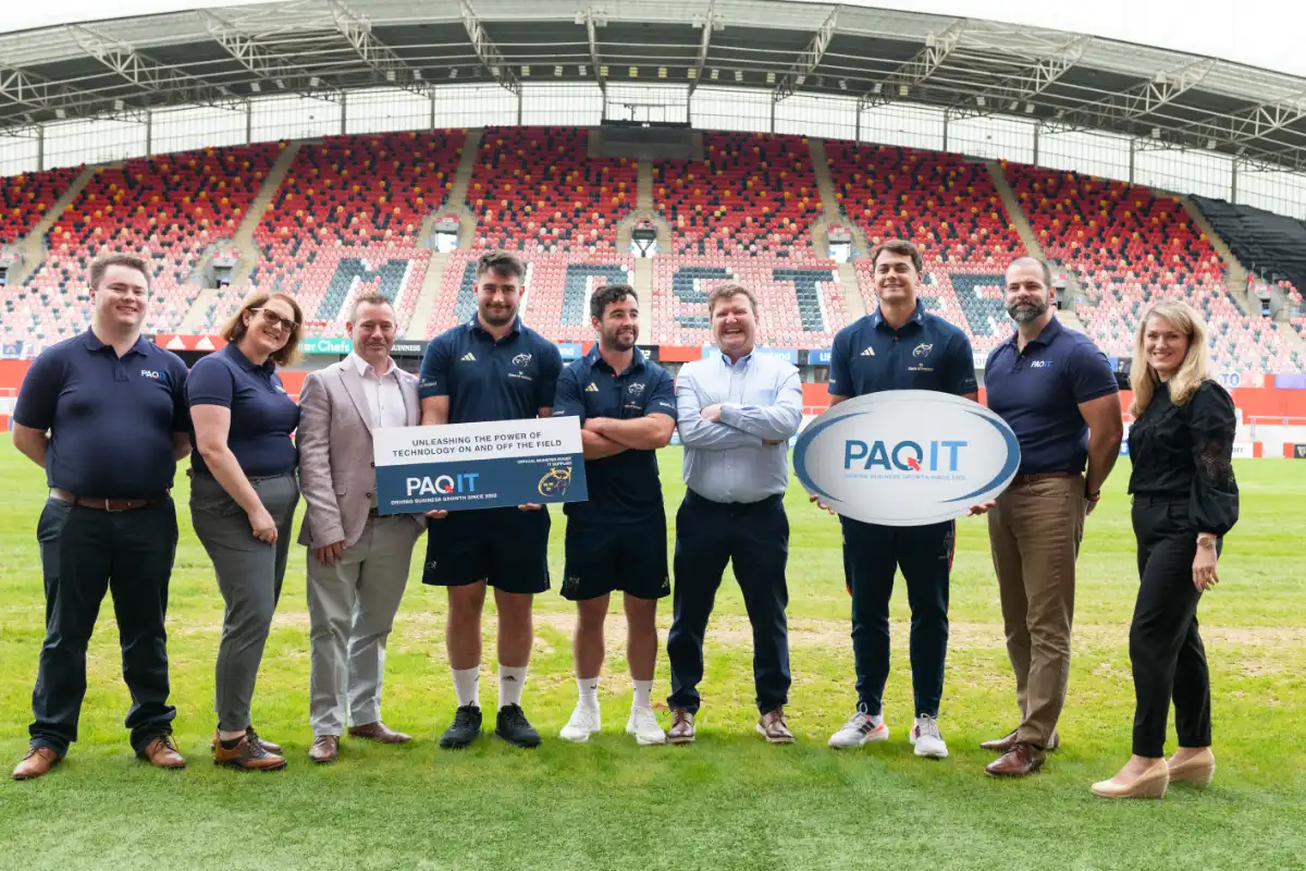 A group of people standing on a rugby field at a stadium, posing for a photo. Some individuals are wearing PAQ IT-branded polo shirts, while others are in Munster Rugby team uniforms.